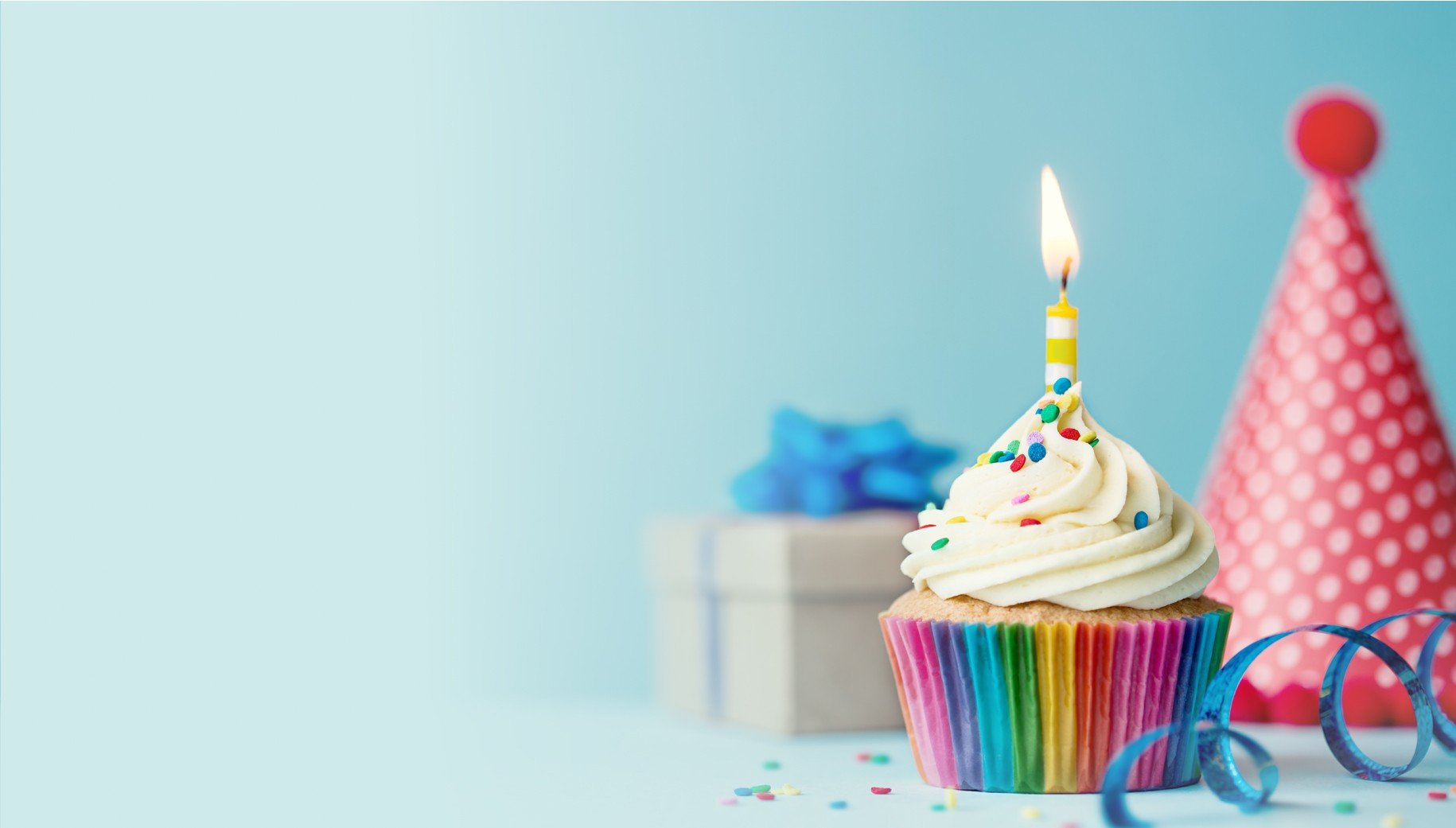 Banner image for online birthday cards, showing cupcake with candle, birthday hat, and present on light blue background